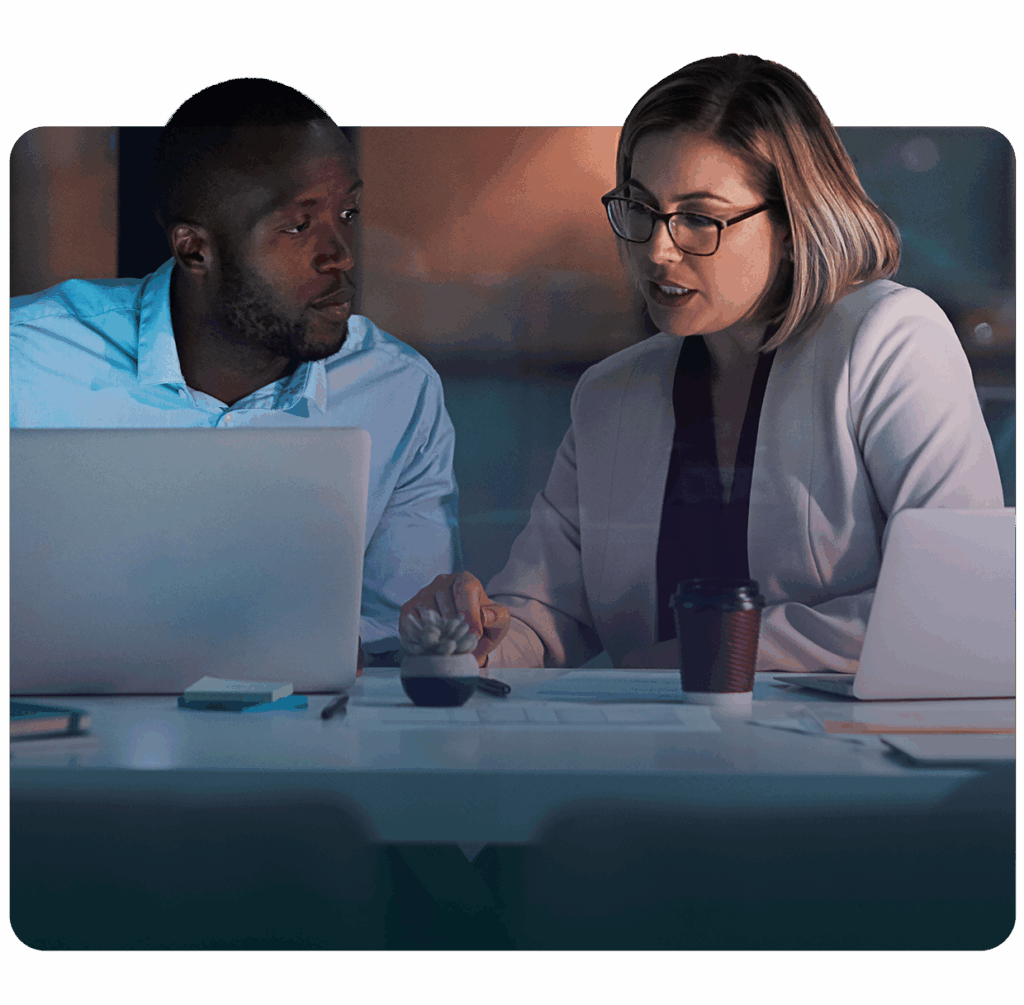 A man and woman are sitting at a workspace reviewing reports on paper and on a laptop