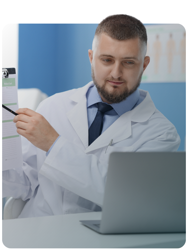 Male clinician looking at laptop while holding up paper and pointing to it