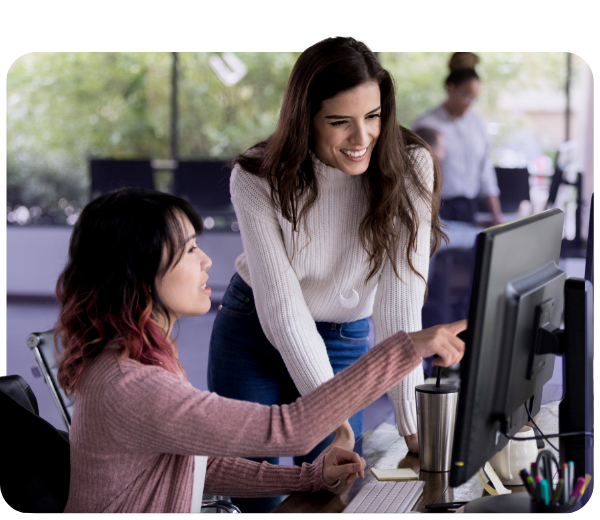 A standing woman and a seated woman pointing at monitor both smiling